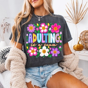 Op de afbeelding: Donkergrijs t-shirt met de tekst "ADULTING is a scam" in kleurrijke letters, omringd door levendige bloemenontwerpen. Een casual kledingstuk.