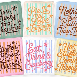 May include: Six folded t-shirts in various pastel colors, each featuring vertical stripes and star accents. The shirts display phrases including Most Likely to be Thankful, Pie Is my Love Language, Nothing Gets Butter Than This, Stuffed Blessed & Thankful, Eat, Drink & Cranberry, and Hot Stuffing Coming Through.