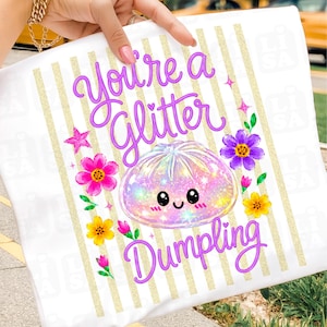 Może przedstawiać: Biała koszulka z błyszczącą grafiką pierożka i fioletowym napisem "You're a Glitter Dumpling". Projekt zawiera kolorowe kwiaty i gwiazdki na tle złotych pionowych pasków.