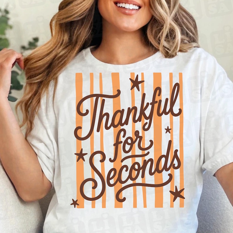 May include: A white short-sleeved t-shirt featuring a graphic design with vertical orange stripes. The text "Thankful for Seconds" is written in a dark brown cursive font, accented by several small dark brown star shapes scattered around.