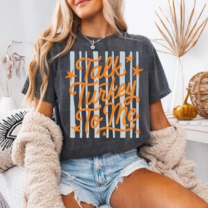 May include: A charcoal gray short-sleeve t-shirt featuring a graphic print with vertical light blue stripes and the orange text "Talk Turkey To Me" accented with small orange stars. The shirt has a crew neckline and a relaxed fit.