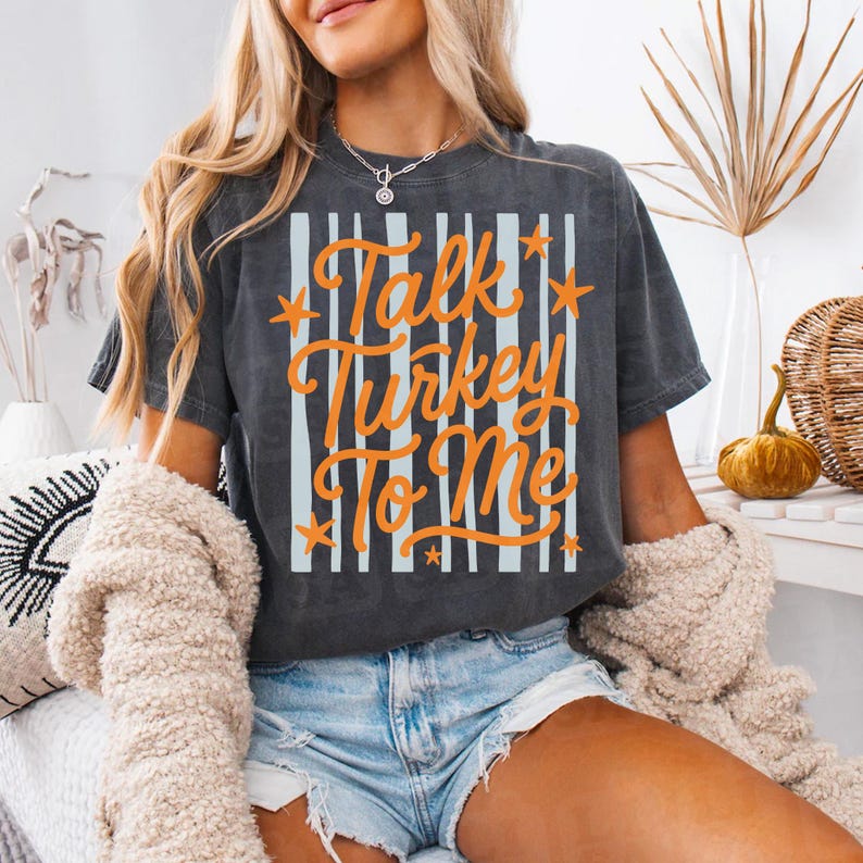 May include: A charcoal gray short-sleeve t-shirt featuring a graphic print with vertical light blue stripes and the orange text "Talk Turkey To Me" accented with small orange stars. The shirt has a crew neckline and a relaxed fit.