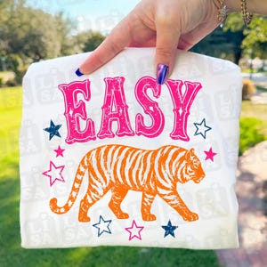Może przedstawiać: Biała koszulka z napisem "EASY" w kolorze różowym i pomarańczowym, ilustracją tygrysa i gwiazdkami. Tygrys jest pomarańczowy z czarnymi paskami. Gwiazdy są różowe i niebieskie.