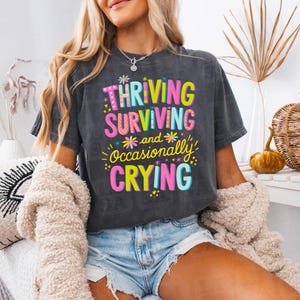 May include: Dark gray t-shirt with the text "Thriving, Surviving, and Occasionally Crying" in colorful, playful lettering. The design includes floral and star accents. The shirt is a casual, comfortable style.