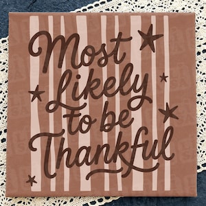 May include: A square sign with a terracotta background featuring vertical light pink stripes. The dark brown text reads Most Likely to be Thankful in a cursive script. Five dark brown stars of varying sizes are positioned around the lettering.