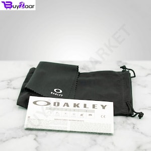 K&ouml;nnte beinhalten: Ein schwarzes Oakley Mikrofasertuch und ein Kordelzugbeutel mit dem Oakley-Logo. Eine wei&szlig;e Karte mit dem Oakley-Logo und Text ist ebenfalls vorhanden. Die Artikel befinden sich auf einer wei&szlig;en Marmoroberfl&auml;che.