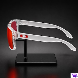 K&ouml;nnte beinhalten: Transparente Sonnenbrille mit roten Gl&auml;sern wird auf einem schwarzen St&auml;nder pr&auml;sentiert. Die Sonnenbrille hat eine rechteckige Form und wei&szlig;e B&uuml;gel. Das Oakley-Logo ist auf dem Rahmen sichtbar. Der Hintergrund ist schwarz.