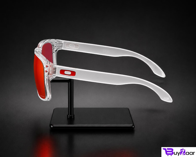 K&ouml;nnte beinhalten: Transparente Sonnenbrille mit roten Gl&auml;sern wird auf einem schwarzen St&auml;nder pr&auml;sentiert. Die Sonnenbrille hat eine rechteckige Form und wei&szlig;e B&uuml;gel. Das Oakley-Logo ist auf dem Rahmen sichtbar. Der Hintergrund ist schwarz.
