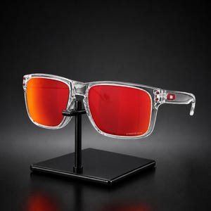 K&ouml;nnte beinhalten: Eine Sonnenbrille mit transparentem Rahmen und leuchtend roten Gl&auml;sern wird auf einem schwarzen St&auml;nder pr&auml;sentiert. Die rechteckigen Gl&auml;ser haben eine leichte Kr&uuml;mmung, und der Rahmen ist transparent. Der Hintergrund ist dunkel.