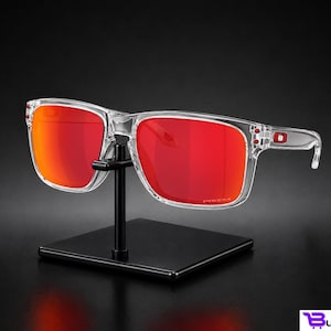 K&ouml;nnte beinhalten: Transparente Sonnenbrille mit leuchtend roten Gl&auml;sern wird auf einem schwarzen St&auml;nder pr&auml;sentiert. Die Sonnenbrille hat eine quadratische Form und ein modernes Design. Die Gl&auml;ser sind leuchtend rot und der Rahmen ist transparent.