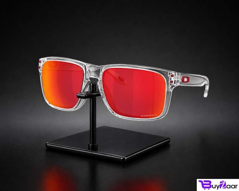 K&ouml;nnte beinhalten: Transparente Sonnenbrille mit leuchtend roten Gl&auml;sern wird auf einem schwarzen St&auml;nder pr&auml;sentiert. Die Sonnenbrille hat eine quadratische Form und ein modernes Design. Die Gl&auml;ser sind leuchtend rot und der Rahmen ist transparent.