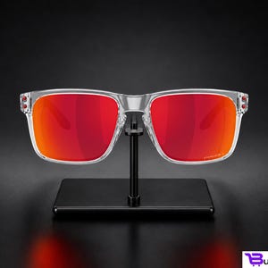 K&ouml;nnte beinhalten: Sonnenbrille mit transparentem Rahmen und leuchtend roten Gl&auml;sern. Die Sonnenbrille wird auf einem schwarzen St&auml;nder vor dunklem Hintergrund pr&auml;sentiert. Die Gl&auml;ser weisen einen Farbverlauf von tiefem Rot zu einem helleren Orangeton auf.