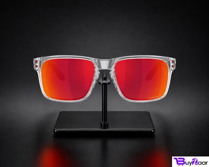 K&ouml;nnte beinhalten: Sonnenbrille mit transparentem Rahmen und leuchtend roten Gl&auml;sern. Die Sonnenbrille wird auf einem schwarzen St&auml;nder vor dunklem Hintergrund pr&auml;sentiert. Die Gl&auml;ser weisen einen Farbverlauf von tiefem Rot zu einem helleren Orangeton auf.