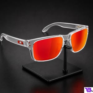 K&ouml;nnte beinhalten: Eine Sonnenbrille mit transparentem Rahmen und leuchtend roten Gl&auml;sern wird auf einem schwarzen St&auml;nder pr&auml;sentiert. Die Sonnenbrille hat eine rechteckige Form und ein kleines rotes Logo am B&uuml;gel. Die Gl&auml;ser reflektieren einen hellen, orange-roten Farbton.