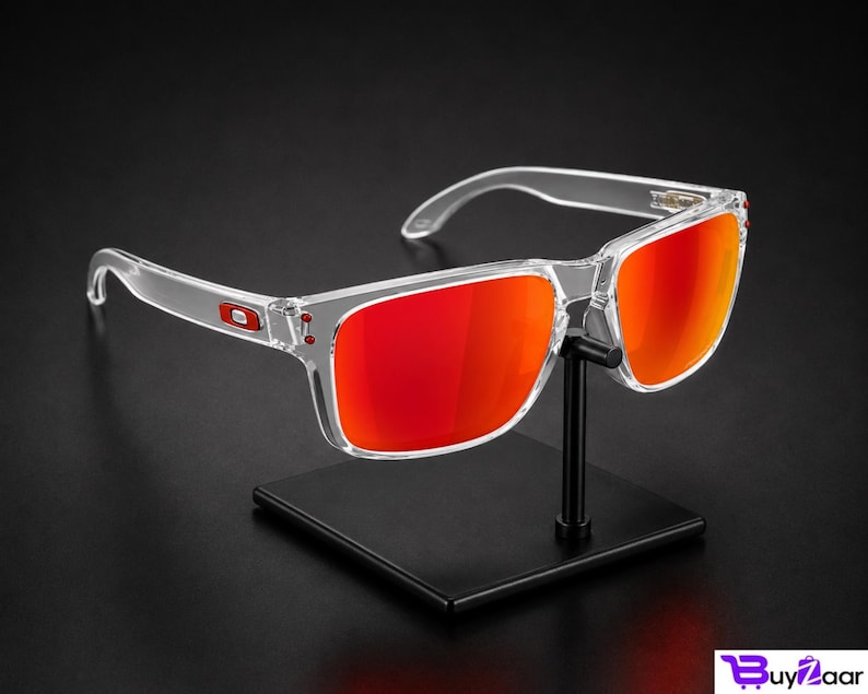 K&ouml;nnte beinhalten: Eine Sonnenbrille mit transparentem Rahmen und leuchtend roten Gl&auml;sern wird auf einem schwarzen St&auml;nder pr&auml;sentiert. Die Sonnenbrille hat eine rechteckige Form und ein kleines rotes Logo am B&uuml;gel. Die Gl&auml;ser reflektieren einen hellen, orange-roten Farbton.