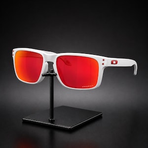Puede incluir: Gafas de sol blancas con lentes rojas sobre un soporte negro. Las gafas tienen una montura rectangular blanca y un logotipo rojo en la patilla. Las lentes son de color rojo brillante.