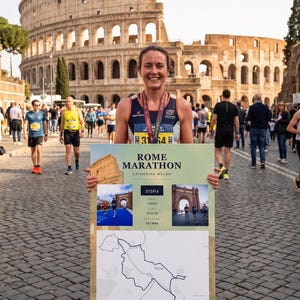 May include: A woman smiles, holding a sign for the Rome Marathon in front of the Colosseum. The sign includes the text "Rome Marathon," a map of the course, and race details. The woman wears a race bib and medal, with other runners visible.