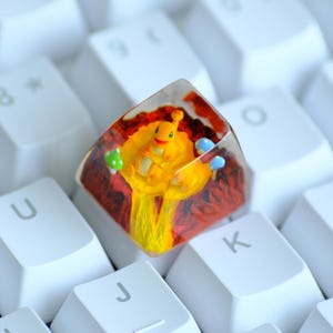 May include: A custom artisan keycap featuring a yellow character with a red and orange background. The keycap is made of clear resin and sits on a white keyboard. The character is holding a green and blue object.