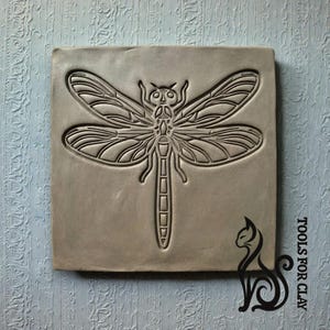 Puede incluir: Una baldosa cuadrada de arcilla gris con un diseño de libélula en relieve. Las alas, el cuerpo y la cabeza de la libélula están claramente definidos. La baldosa está sobre un fondo azul claro texturizado. Una silueta de gato negro y el texto "TOOLS FOR CLAY" están en la esquina inferior derecha.