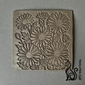 Puede incluir: Un sello cuadrado de arcilla gris con un relieve detallado de margaritas y follaje. El diseño incluye varias margaritas grandes con pétalos y hojas detallados, creando un patrón floral. El sello es probablemente para usar en arte de arcilla.