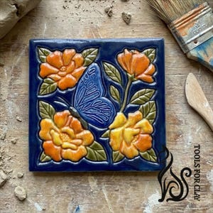 Puede incluir: Azulejo de cerámica cuadrado con un diseño en relieve que presenta una mariposa azul y flores naranjas y amarillas sobre un fondo azul oscuro. Las flores tienen hojas verdes. El azulejo está sobre una superficie de madera.