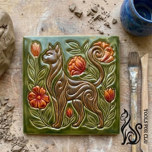 Puede incluir: Azulejo cerámico cuadrado con un diseño de gato marrón rodeado de flores naranjas y hojas verdes. El azulejo tiene un acabado brillante. El azulejo mide aproximadamente 15 cm por 15 cm. La imagen también muestra herramientas de cerámica.