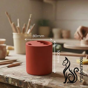 May include: A cylindrical, terracotta-colored clay tool holder, 100mm tall and 80mm wide, with a wavy top. The image includes the text "TOOLS FOR CLAY" and a cat silhouette. Paintbrushes and other tools are visible in the background.