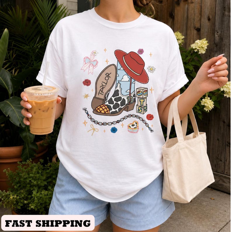 Op de afbeelding: Een wit t-shirt met een grafische print van een cowboylaars met koeienprint, een rode hoed, de naam Taylor en blokken met de letters TS. Het ontwerp bevat kleine bloemen, een strik en een kralensnoer rondom de centrale laarsillustratie.