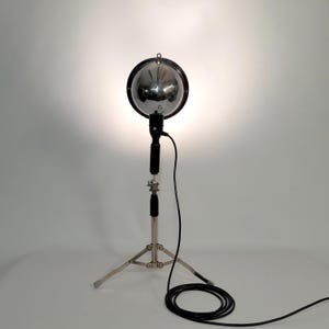 Könnte beinhalten: Ein Vintage-Chrom-Scheinwerfer mit einem kugelförmigen Reflektor und einem schwarzen Griff, montiert auf einem Stativ. Der Scheinwerfer hat einen kreisförmigen Rahmen und ein schwarzes Netzkabel. Das Licht scheint hell.