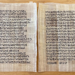 May include: Two parchment scrolls with ancient Greek text in black ink. The text is divided into two columns, with each scroll containing several lines of script. The paper has a beige hue, and the text appears to be a religious passage.