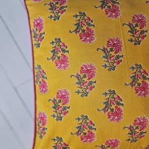 Könnte beinhalten: Senfgelbes Zierkissen mit floralem Muster. Das Kissen hat ein sich wiederholendes Design aus rosa Blüten und grünen Blättern. Die Kanten sind mit einem dünnen rosa Rand versehen.