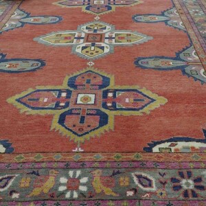 May include: A large, rectangular rug with a terracotta background and intricate geometric patterns in blue, yellow, pink, and gray. The rug has a detailed border and fringe along the edges. A woven basket is in the upper left corner.