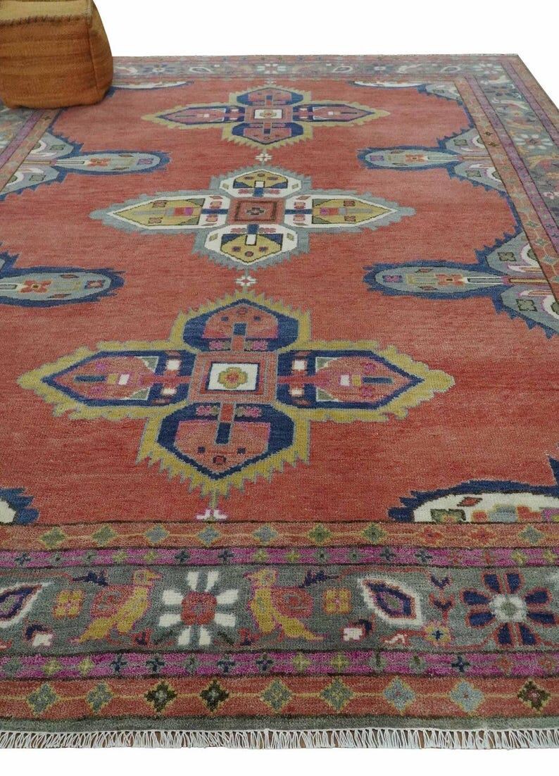 May include: A large, rectangular rug with a terracotta background and intricate geometric patterns in blue, yellow, pink, and gray. The rug has a detailed border and fringe along the edges. A woven basket is in the upper left corner.