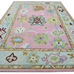 May include: A rectangular pink rug with a floral design. The rug features a light blue border with floral and geometric patterns. The central design includes various floral motifs in shades of green, brown, and blue. The rug is a decorative home furnishing.