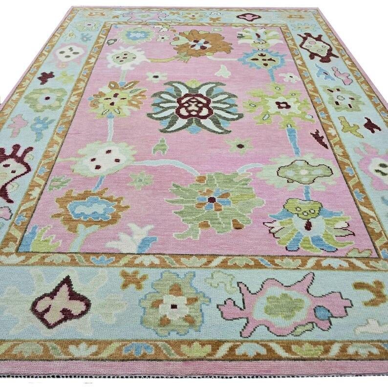 May include: A rectangular pink rug with a floral design. The rug features a light blue border with floral and geometric patterns. The central design includes various floral motifs in shades of green, brown, and blue. The rug is a decorative home furnishing.