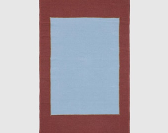 Op de afbeelding: Een rechthoekig vloerkleed met een lichtblauw midden en een donkerrode rand. Een dunne goudkleurige lijn scheidt de blauwe en rode delen. Het vloerkleed heeft een eenvoudig, modern ontwerp.