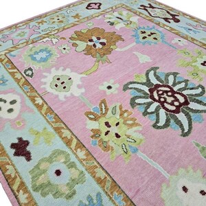 May include: A pink area rug with a floral pattern. The rug features large, stylized flowers in shades of brown, green, and blue against a pink background. The border is a light blue with a repeating floral design. The rug is rectangular.