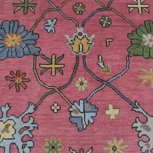 May include: Close-up of a pink rug with a detailed floral pattern. The design includes large blue, green, and yellow flowers, plus smaller floral and geometric shapes in various colours. The rug has a textured appearance.