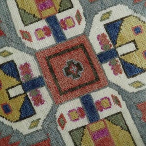 May include: Close-up of a colorful, patterned rug. The rug features a central red square with a cross-like design, surrounded by white, blue, yellow, and gray geometric shapes. The edges are a reddish-orange color.