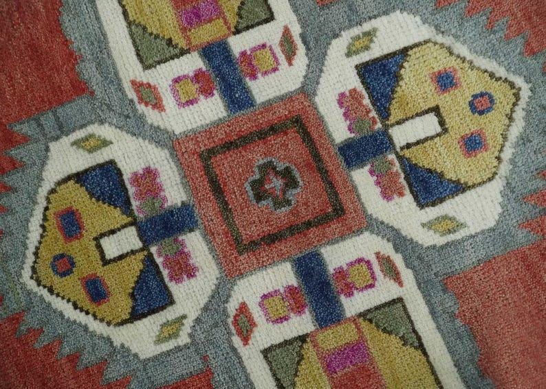 May include: Close-up of a colorful, patterned rug. The rug features a central red square with a cross-like design, surrounded by white, blue, yellow, and gray geometric shapes. The edges are a reddish-orange color.