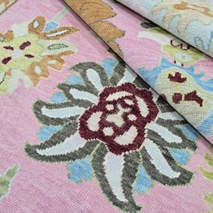 May include: A close-up of a pink rug with a floral pattern. The rug features large, stylized flowers in shades of burgundy, gray, blue, and yellow. The rug is folded over, revealing the underside with a similar pattern. The rug is a decorative home decor item.