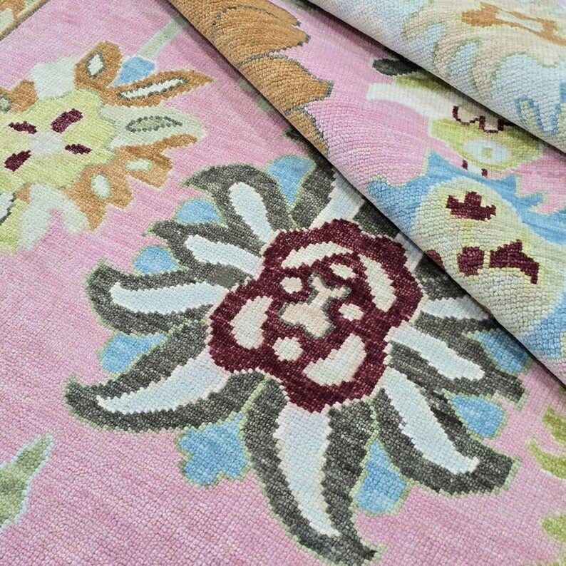 May include: A close-up of a pink rug with a floral pattern. The rug features large, stylized flowers in shades of burgundy, gray, blue, and yellow. The rug is folded over, revealing the underside with a similar pattern. The rug is a decorative home decor item.