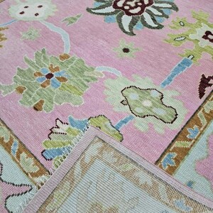 May include: A pink area rug with a floral pattern in shades of green, blue, and brown. The rug has a detailed border and a soft, textured appearance. The design features stylized flowers and leaves.