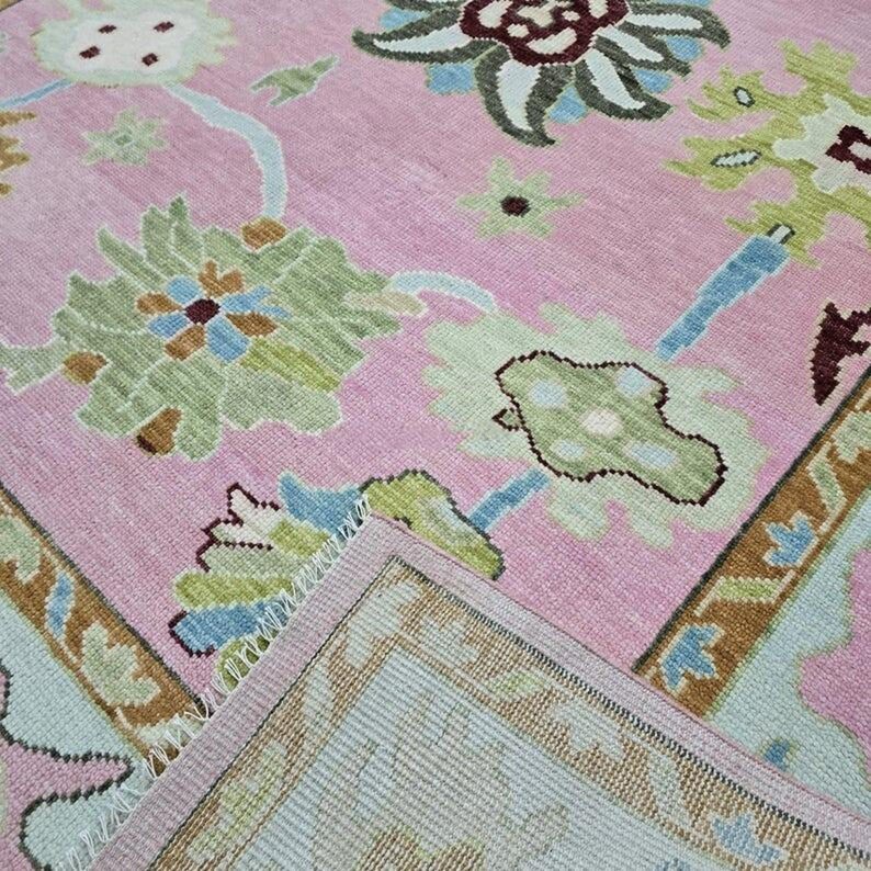 May include: A pink area rug with a floral pattern in shades of green, blue, and brown. The rug has a detailed border and a soft, textured appearance. The design features stylized flowers and leaves.
