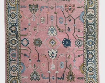 May include: A rectangular pink rug with an intricate floral pattern. The rug features a detailed border in shades of blue, green, and gold. The central design includes stylized flowers and geometric shapes, creating a vintage aesthetic.