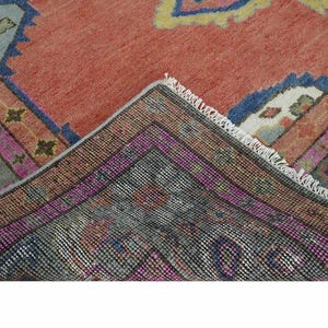 May include: A section of a vintage rug with a warm, reddish-orange central field. The rug features intricate geometric patterns in shades of blue, pink, and green, with a detailed border. The rug's edges are frayed, showing its age and use.