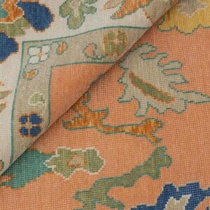 May include: Close-up of a section of a woven rug with a detailed floral pattern. The rug features a peach-coloured background with accents of blue, green, and orange. The border is a light beige with green and orange floral designs.