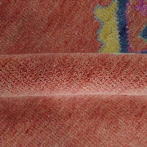 May include: Close-up of a textured, coral-colored rug with a folded section. The rug features geometric patterns in blue, yellow, and purple. The plush pile creates a soft, inviting appearance. This rug is suitable for home decor.