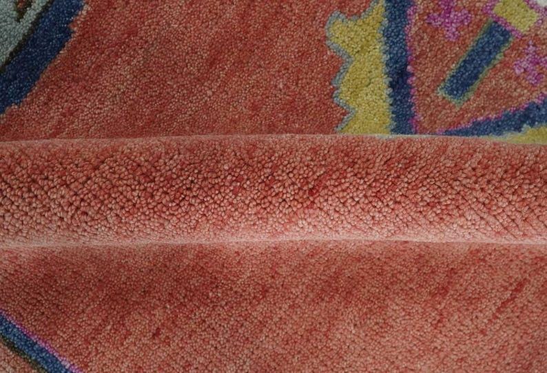 May include: Close-up of a textured, coral-colored rug with a folded section. The rug features geometric patterns in blue, yellow, and purple. The plush pile creates a soft, inviting appearance. This rug is suitable for home decor.