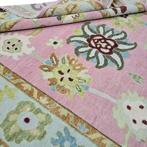 May include: A pink area rug with a floral pattern. The rug features large, stylized flowers in shades of green, blue, and brown against a pink background. The border has a geometric design with similar colors. The rug is partially rolled up.